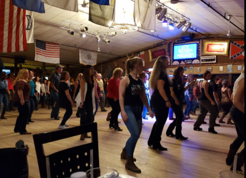 A lively dance floor filled with people line dancing, with flags and lights in the background.