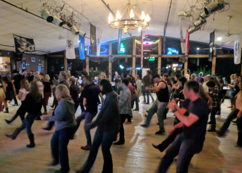 A lively dance floor filled with people line dancing in a spacious venue with colorful lights and decorations.
