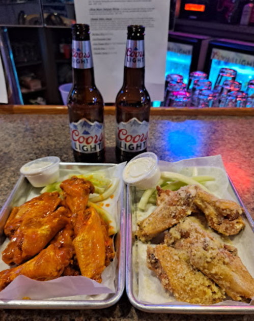 Two trays of chicken wings with dipping sauces, accompanied by two bottles of Coors Light beer on a bar countertop.