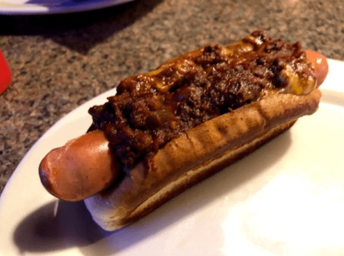 A hot dog topped with chili and cheese, served in a soft bun on a white plate.