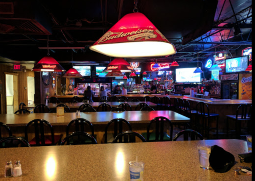 A dimly lit bar with red pendant lights, empty tables, and a long counter lined with drinks and neon signs.