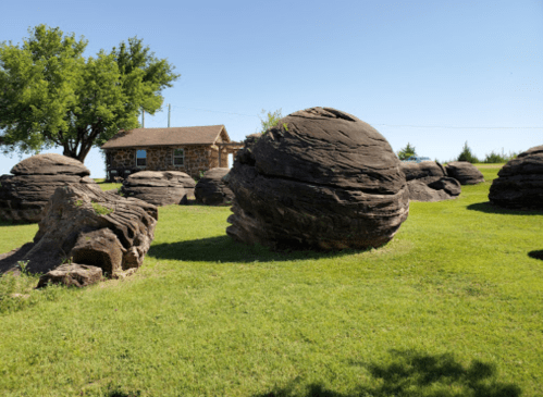 Large, round rock formations scattered across a grassy area, with a small stone house in the background under a clear blue sky.