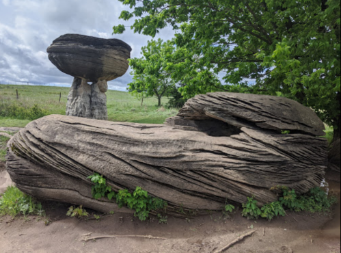 Two large, weathered rock formations with unique textures, surrounded by greenery under a cloudy sky.