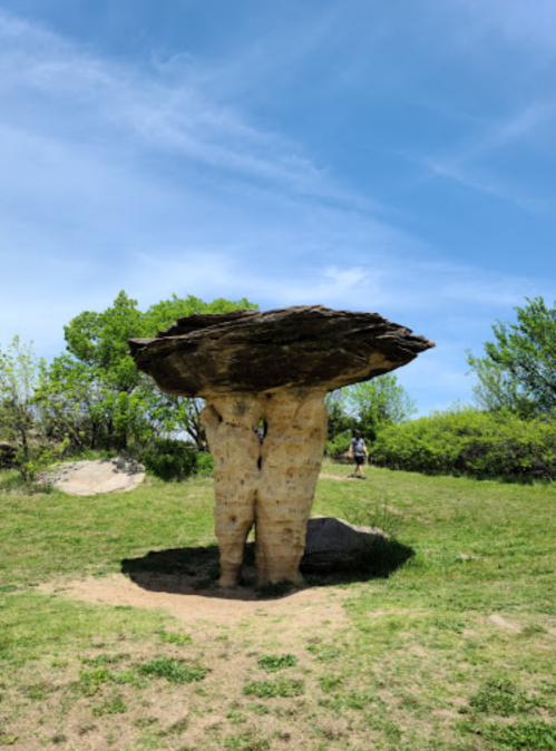 A large rock formation resembling a mushroom, with a wide cap and a narrow base, surrounded by greenery and blue sky.