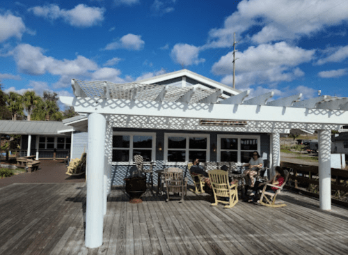 A waterfront restaurant with a wooden deck, featuring rocking chairs and diners enjoying the outdoor seating under a blue sky.