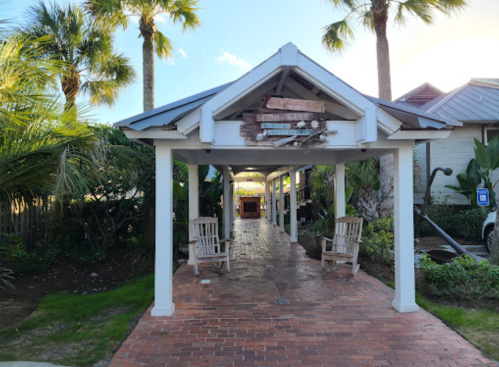A pathway with rocking chairs leads to a building, surrounded by palm trees and lush greenery.