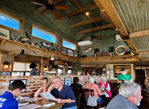 A bustling restaurant interior with wooden beams, brick walls, and patrons dining at tables. Decor includes fishing-themed items.