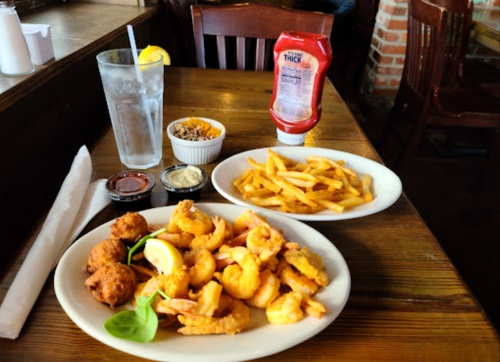A plate of fried shrimp with lemon, hushpuppies, fries, and dipping sauces, alongside a glass of water with lemon.