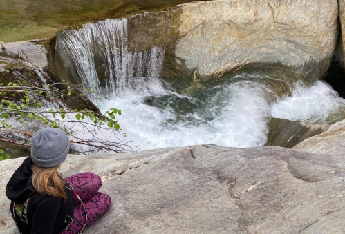 A person sits on a rock by a flowing stream, surrounded by nature and a small waterfall in the background.