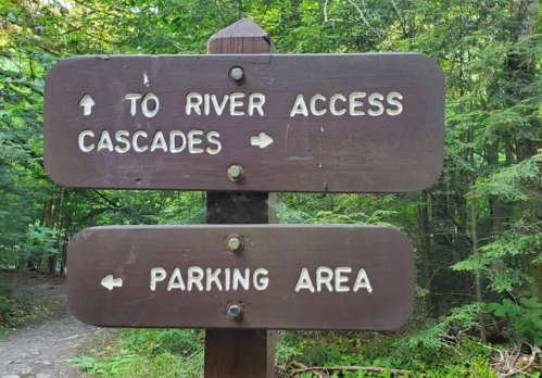 Wooden signpost with arrows indicating directions to river access, cascades, and parking area in a forested setting.