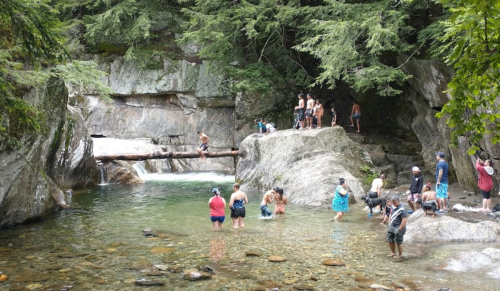 A group of people enjoying a natural swimming area with rocks and trees, some wading in water and others on the shore.