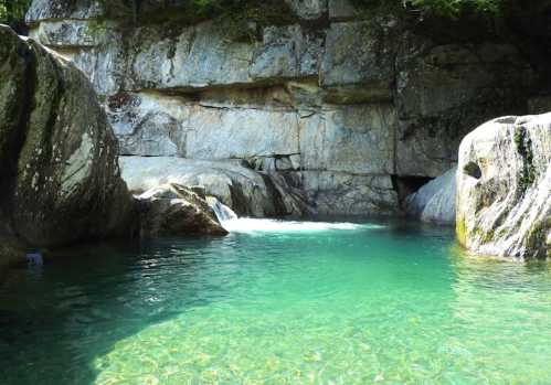 A serene natural pool surrounded by large rocks and a rocky cliff, with clear turquoise water reflecting sunlight.