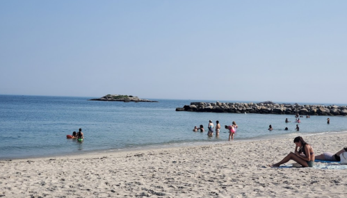 A sunny beach scene with people swimming and relaxing on the sand, with rocky formations in the background.