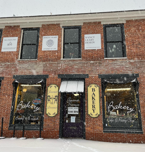 A brick building with a bakery and café, featuring large windows and a snowy exterior, with signs for nearby businesses.
