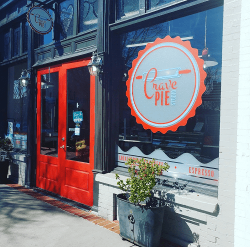Exterior of Crave Pie shop featuring a red door and large window with a logo, plants, and an "Open" sign.