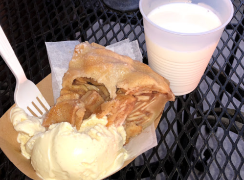 A slice of apple pie with a scoop of vanilla ice cream and a cup of milk on a black metal table.