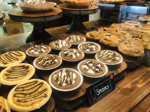 A display of various desserts, including s'mores pies and cookies, arranged on wooden stands in a cozy bakery.