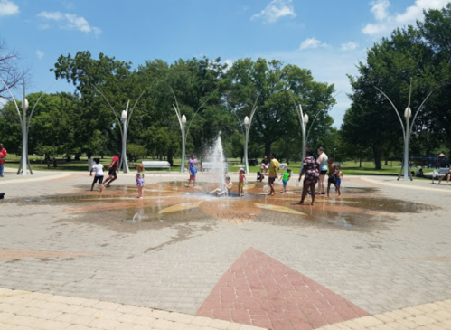 Children and adults play in a splash pad fountain on a sunny day, surrounded by green trees and blue skies.