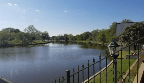 A serene river scene with lush greenery, blue sky, and a decorative fence in the foreground.