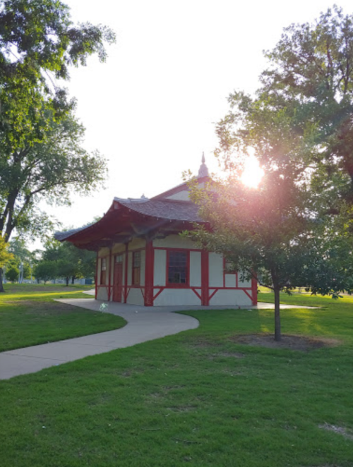 A small, red and white building with a curved roof, surrounded by green grass and trees, with sunlight shining in the background.