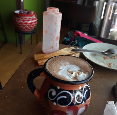 A colorful mug of hot chocolate with whipped cream sits on a table, alongside a partially eaten plate and a bottle.