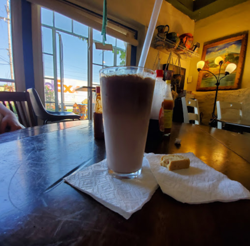 A tall glass of iced coffee on a wooden table, accompanied by a small piece of bread and a napkin.