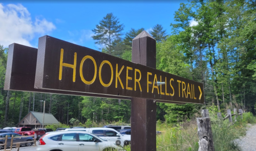 Sign for Hooker Falls Trail with a directional arrow, surrounded by trees and a parking area in the background.