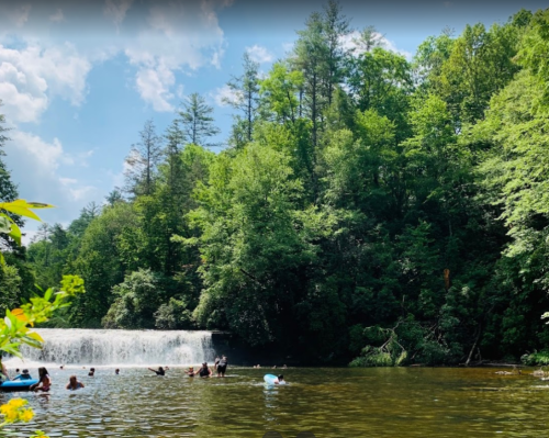 A serene waterfall surrounded by lush greenery, with people enjoying the water on a sunny day.