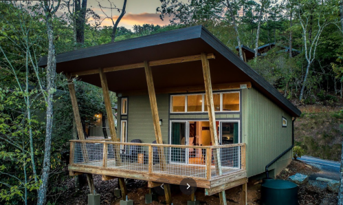 A modern cabin with a sloped roof, large windows, and a wooden deck, surrounded by trees at sunset.