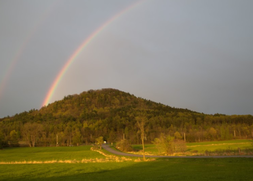 A vibrant rainbow arcs over a green hill, with a winding road and trees in the foreground under a cloudy sky.