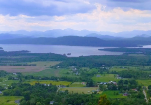 A scenic view of a lake surrounded by green fields and distant mountains under a cloudy sky.