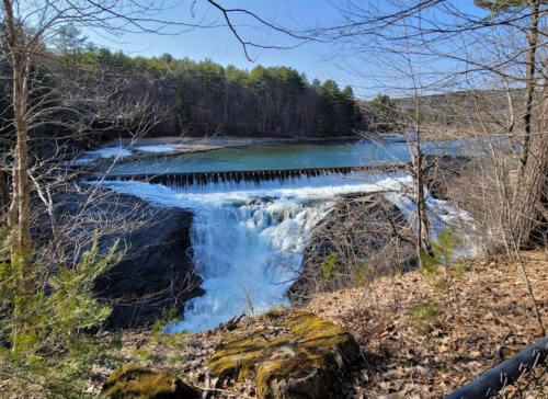 A scenic view of a waterfall cascading over rocks, surrounded by trees and a calm river under a clear blue sky.