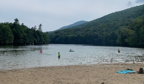 A serene lake surrounded by green mountains, with people swimming and relaxing on the sandy shore.