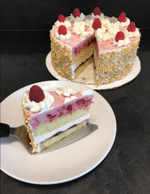 A slice of layered raspberry cake on a plate, with the whole cake decorated with raspberries and cream in the background.