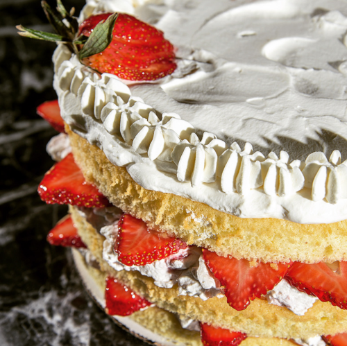 A layered cake with whipped cream and fresh strawberries on top and between the layers, set on a dark marble surface.