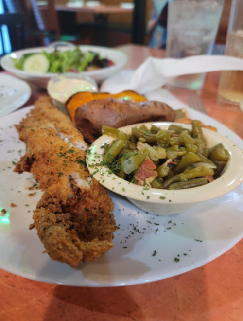 A plate featuring fried fish, green beans, a baked sweet potato, and a side salad with dressings.