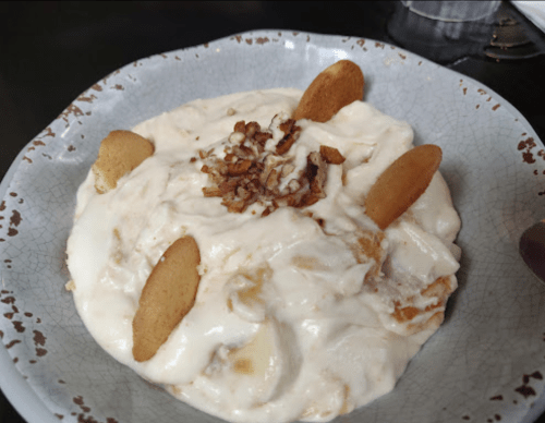 A bowl of creamy dessert topped with crushed nuts and cookie pieces, featuring a smooth, white texture.