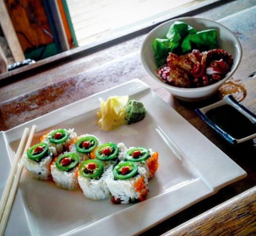 A plate of sushi rolls with green toppings, wasabi, pickled ginger, and a bowl of octopus salad on a wooden table.