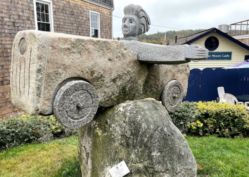 A whimsical stone sculpture of a person in a car, with arms outstretched, set on a large rock in a grassy area.