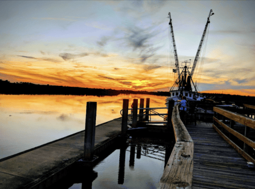 A serene sunset over a calm river, with a fishing boat docked beside a wooden pier.