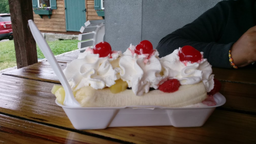 A banana split topped with whipped cream, cherries, and fruit, served in a white dish on a wooden table.