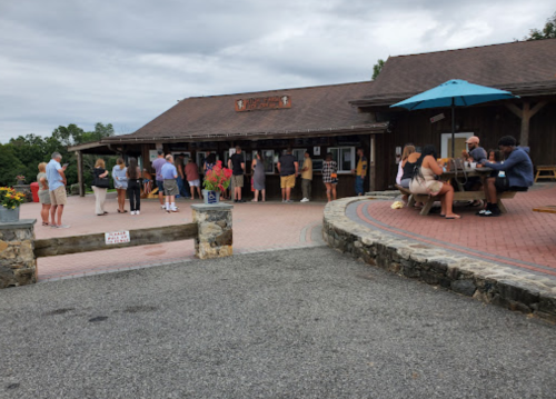 A crowd gathers outside a rustic building with a sign, some people seated at tables under an umbrella.