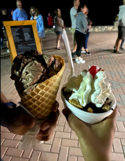 Two hands hold desserts: a chocolate ice cream cone and a sundae topped with whipped cream and a cherry, with people in the background.