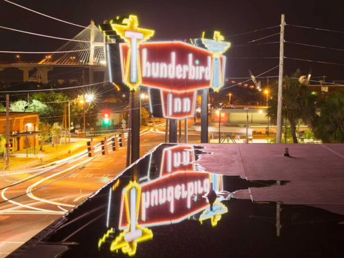 Neon sign for Thunderbird Inn reflected in water at night, with city lights and a bridge in the background.
