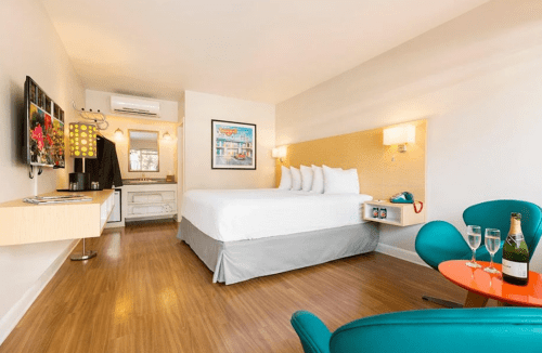 A modern hotel room featuring a queen bed, kitchenette, TV, and a seating area with champagne and glasses.