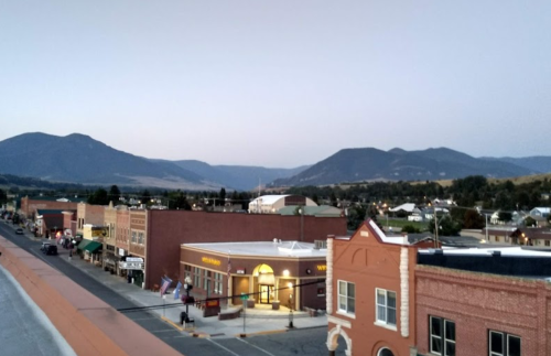 A small town street with brick buildings, shops, and mountains in the background during twilight.