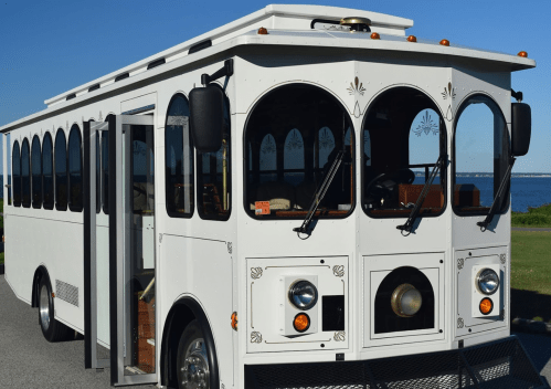 A white trolley bus with large arched windows parked on a scenic road near the water.