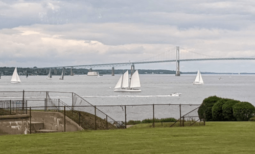 Sailboats on a calm waterway with a bridge in the background under a cloudy sky. Green grass in the foreground.