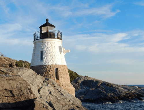 A white lighthouse stands on rocky shores under a blue sky with scattered clouds. Waves gently crash nearby.