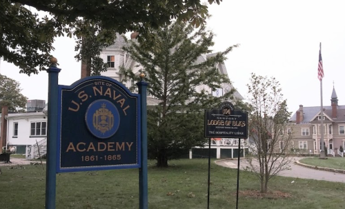 Sign for the U.S. Naval Academy, with a historic building and trees in the background, under a cloudy sky.